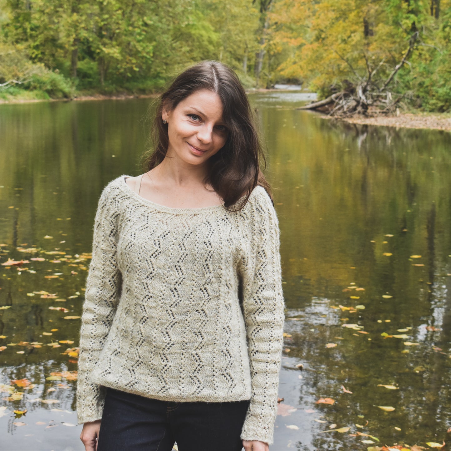 Woman wearing a patterned hand-knit sweater standing by a pond with trees in the background.