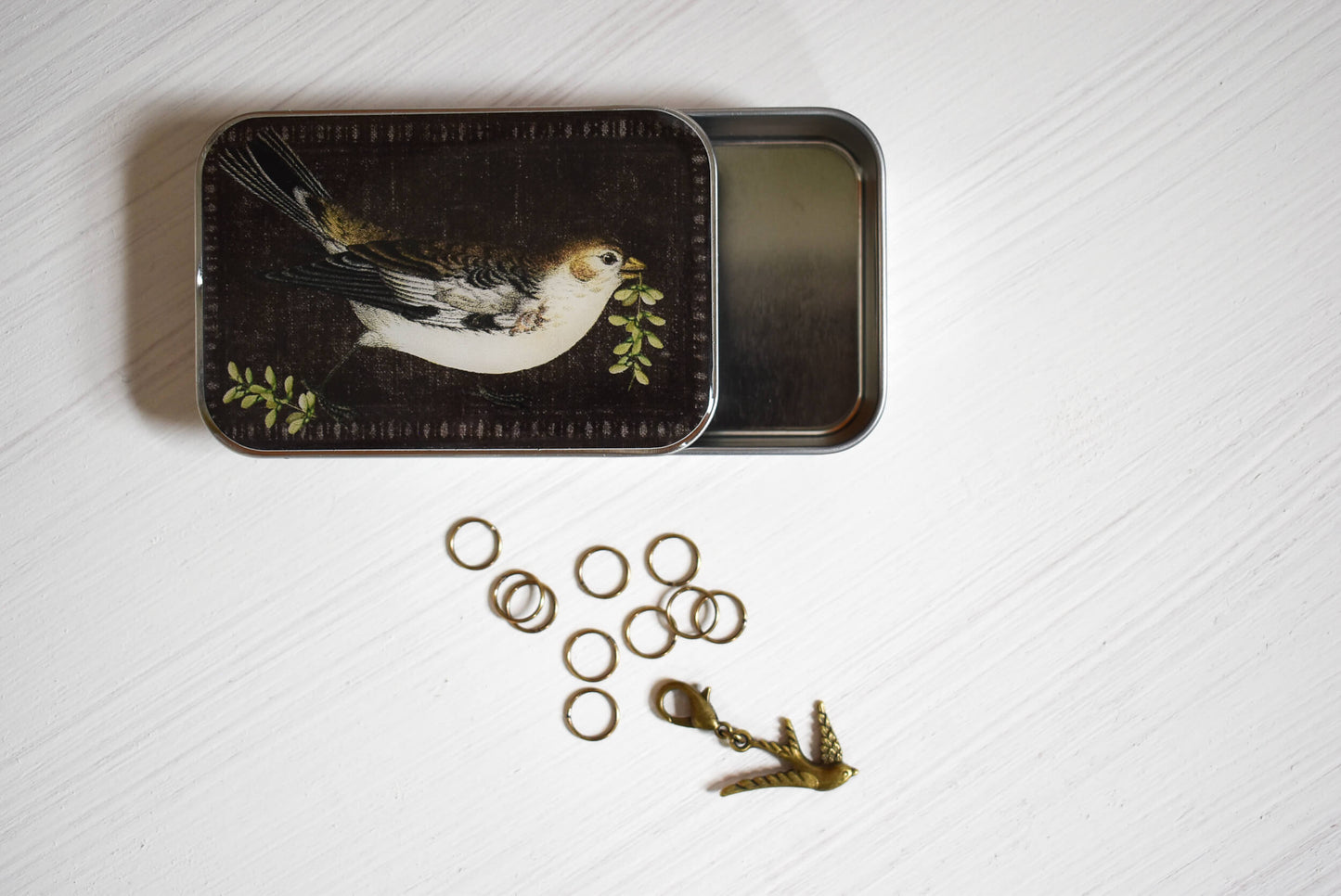 Decorative tin with bird design on lid, surrounded by stitch markers and a bird progress keeper on a textured white surface.