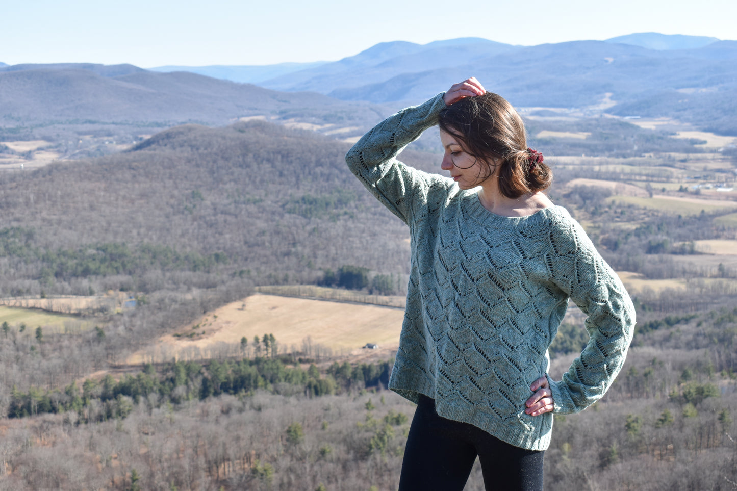 Woman wearing a green Waterscapes sweater standing in a scenic landscape with mountains and fields.