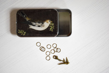 Decorative tin with bird design on lid, surrounded by stitch markers and a bird progress keeper on a textured white surface.