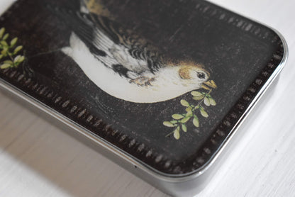 Detail of the knitting notions tin with a bird design on a dark background.