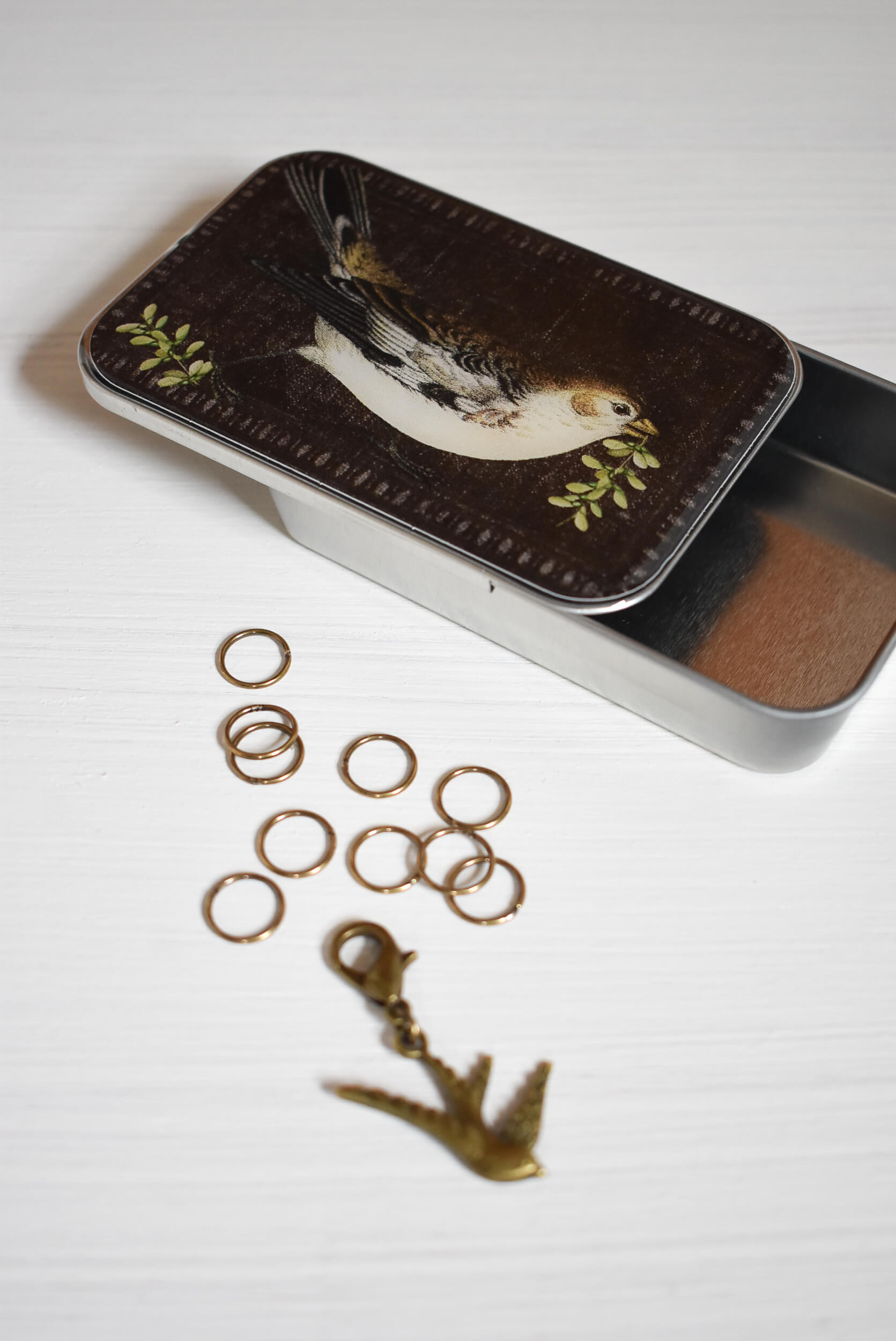Small tin box with bird design on lid, containing gold stitch markers and a bird progress keeper for knitting.