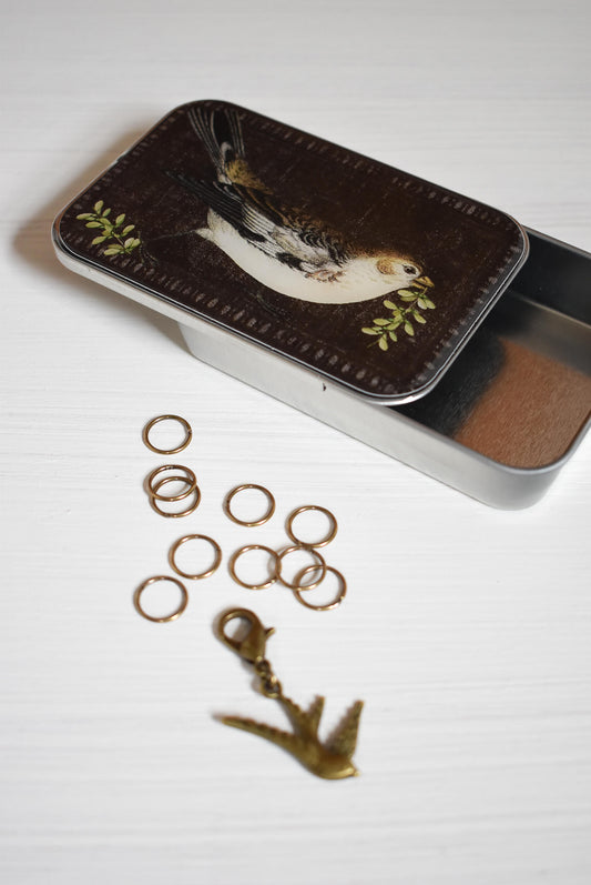 Small tin box with bird design on lid, containing gold stitch markers and a bird progress keeper for knitting.