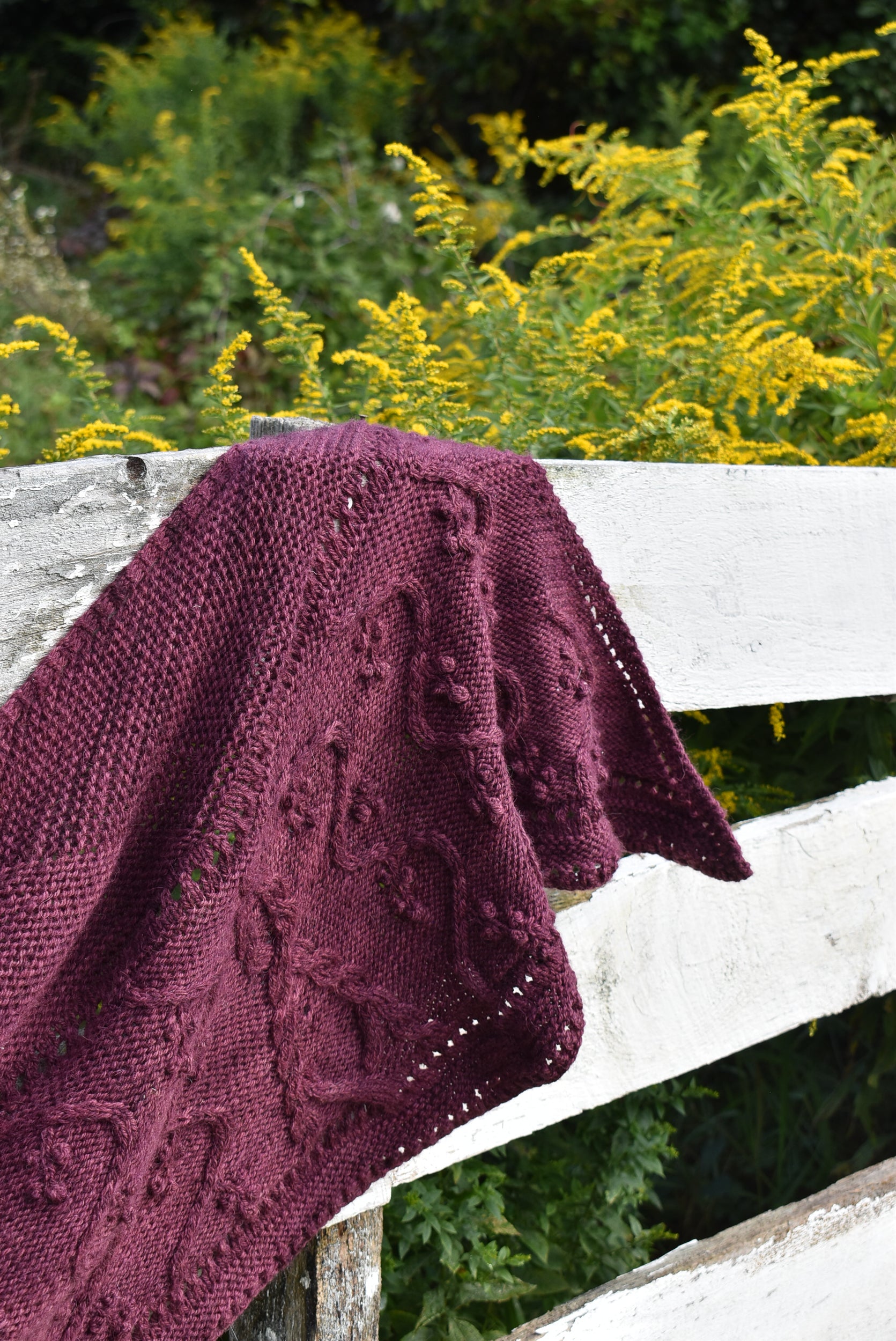 Purple hand-knit shawl on a white wooden fence with greenery in the background, showing the Lady of the Lake knitting pattern.