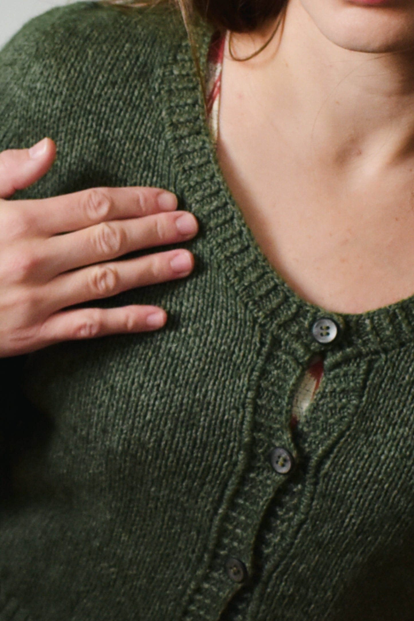 Person wearing a green knitted cardigan with buttons and tuck stitch detail, hand on chest.