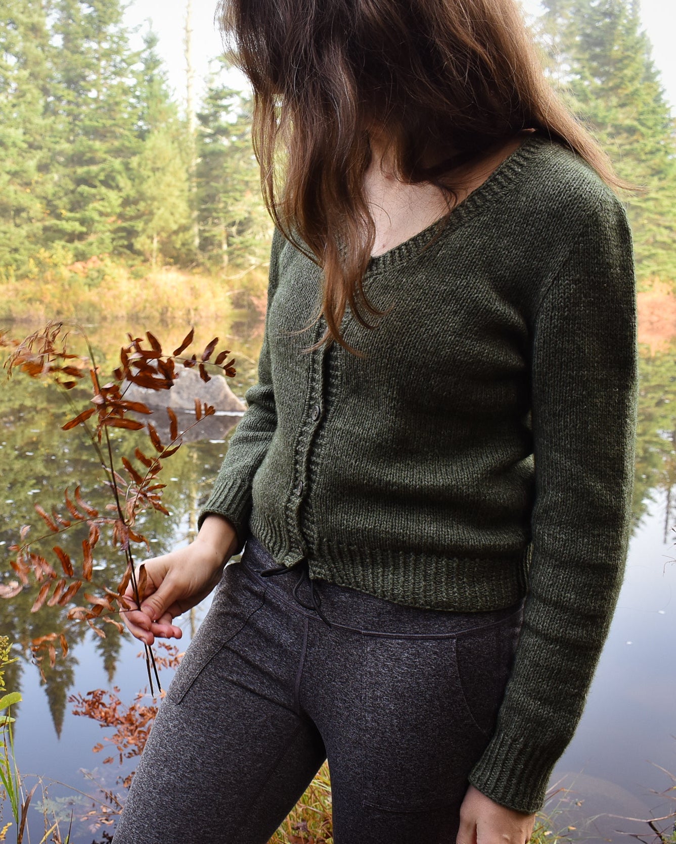 A photo showing a woman wearing the Deepwood Cardigan by a lake with trees in the background.