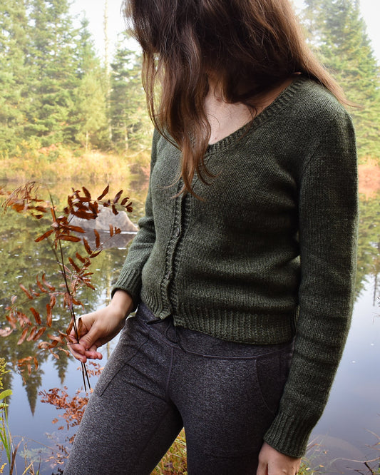 A photo showing a woman wearing the Deepwood Cardigan by a lake with trees in the background.