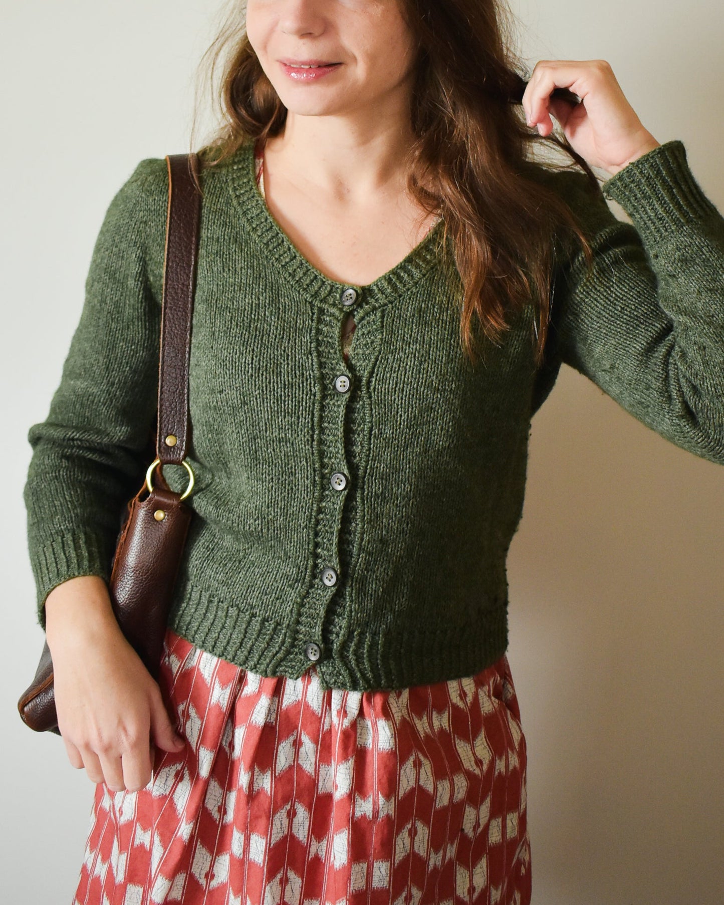 Photo of a woman wearing a green knitted cardigan over a red patterned dress, holding a brown bag.