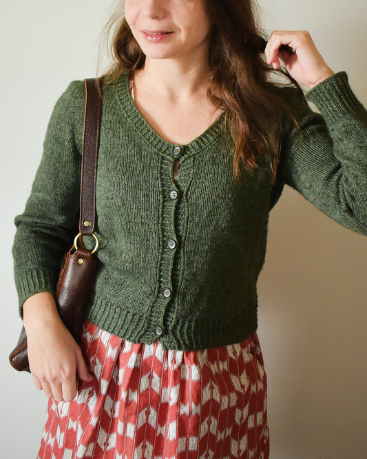 Photo of a woman wearing a green knitted cardigan over a red patterned dress, holding a brown bag.