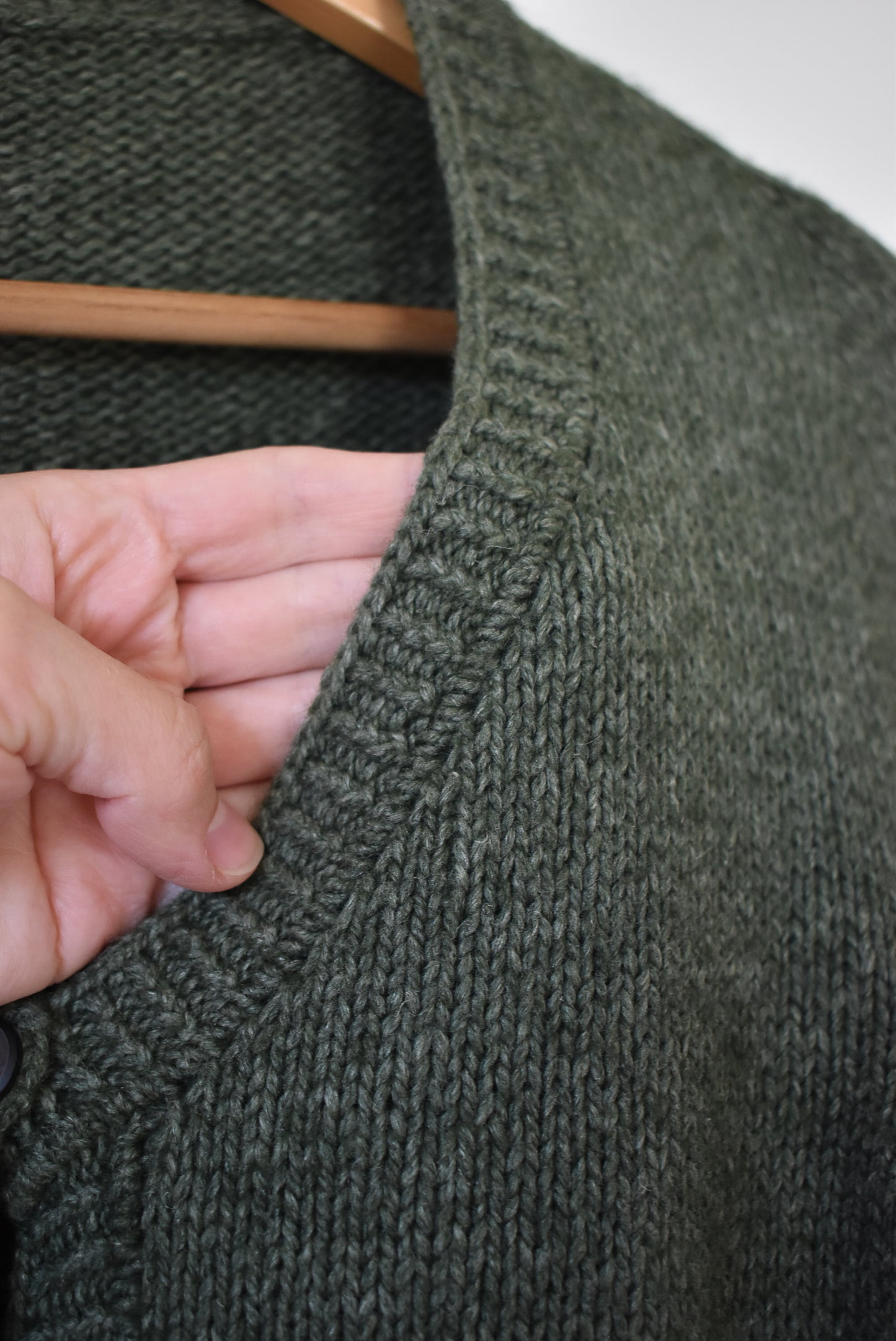 Close-up of a dark green knitted cardigan on a hanger with a hand touching the fabric, showing the tuck stitch detail on the neckband.