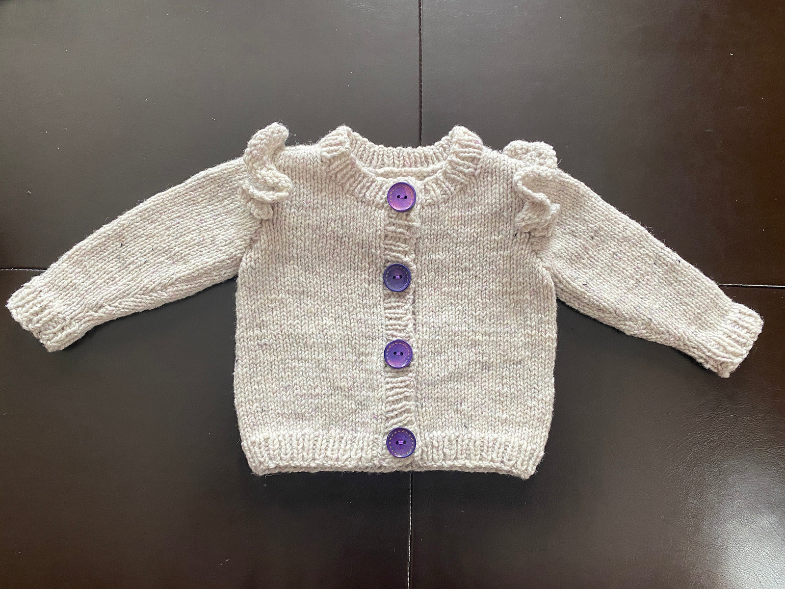 Knitted sweater based on the Ruffle Baby Cardigan knitting pattern with purple buttons on a dark surface.