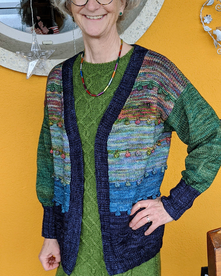 Woman wearing a green dress and colorful cardigan standing in front of a yellow wall with a round mirror.