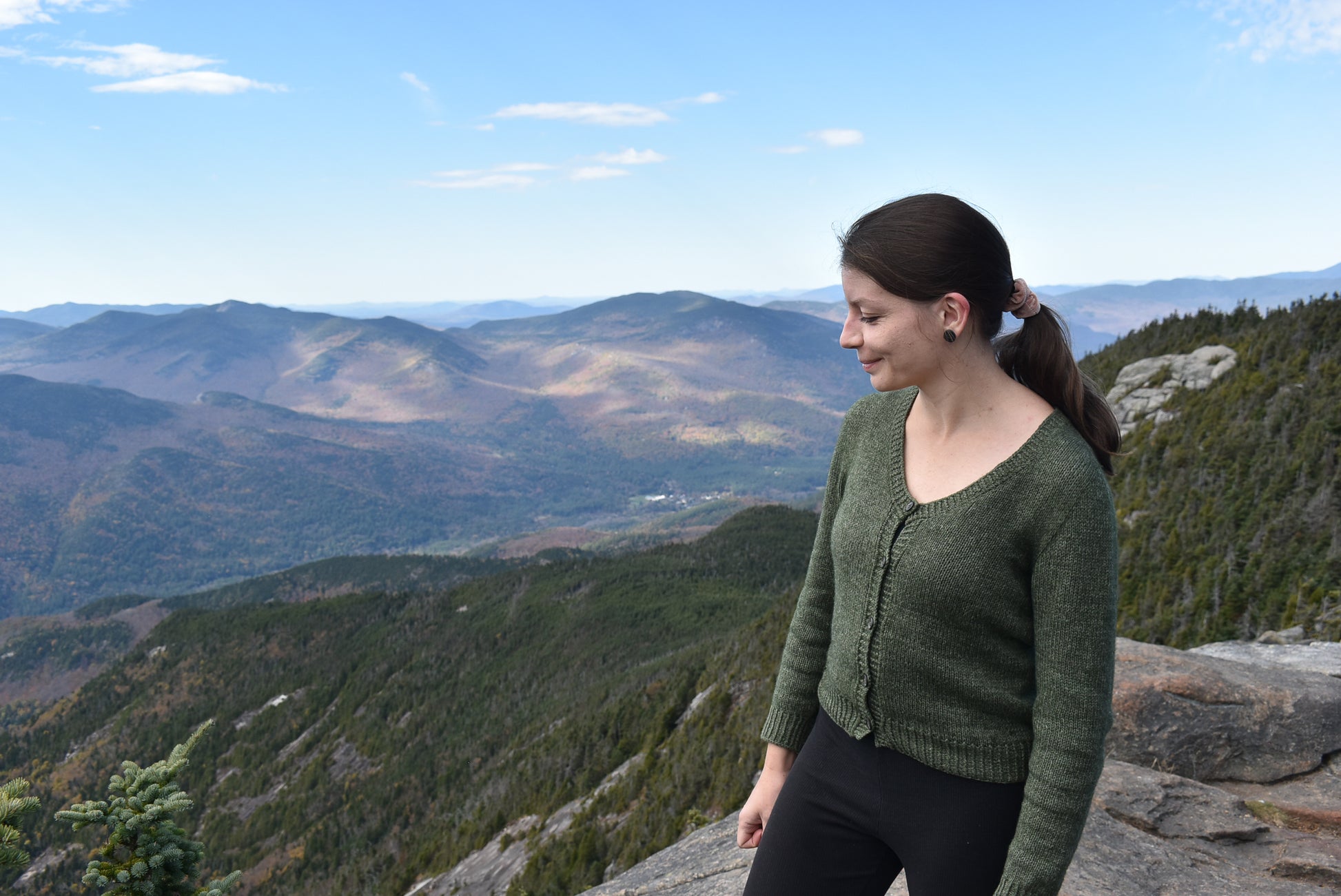 Woman in a green cardigan based on the Deepwood Cardigan knitting pattern, standing on a mountain with a scenic view of mountains and trees.