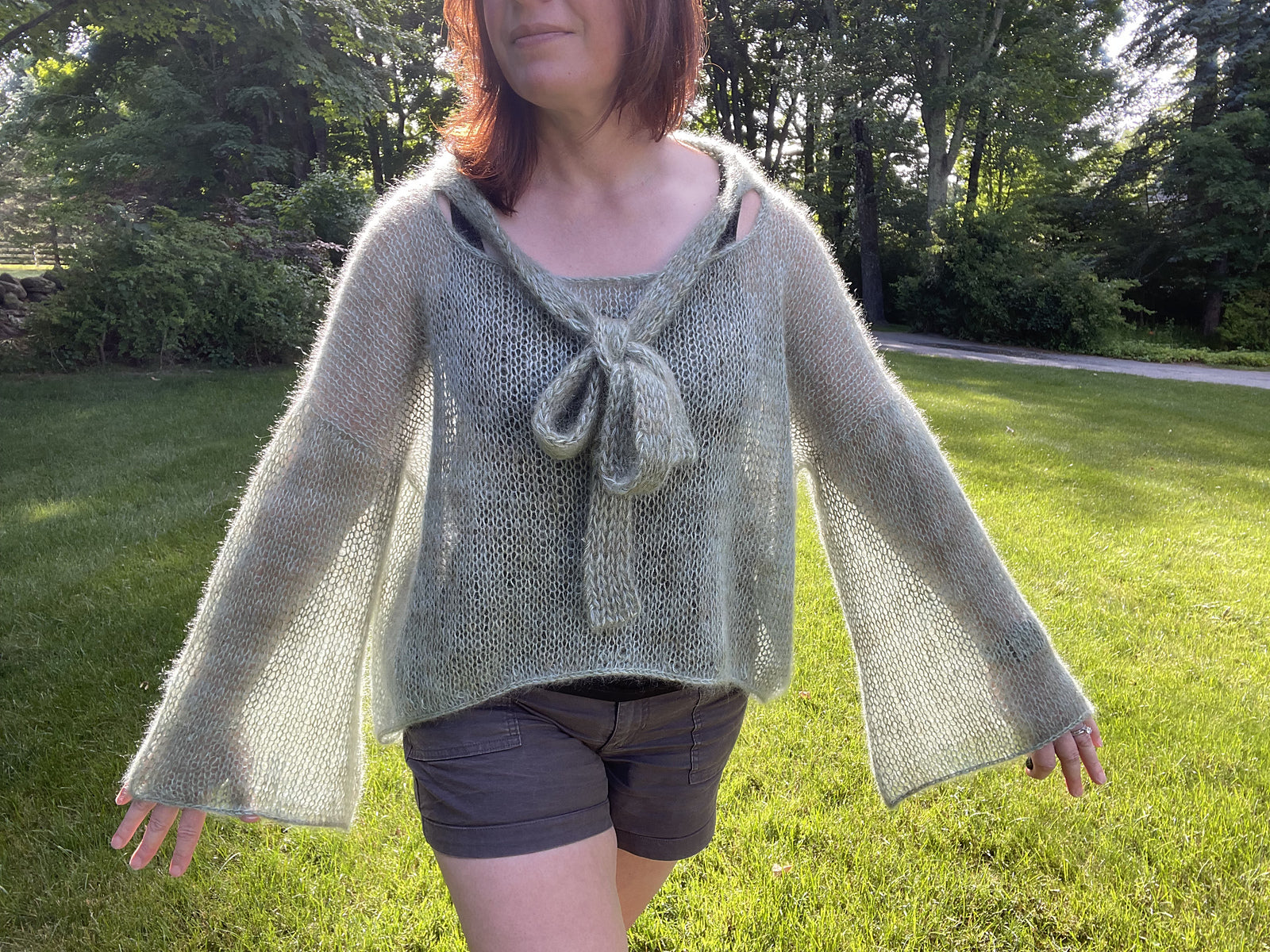 Completed West Village Blouse knitting pattern project in green color, worn outside with trees in the background.