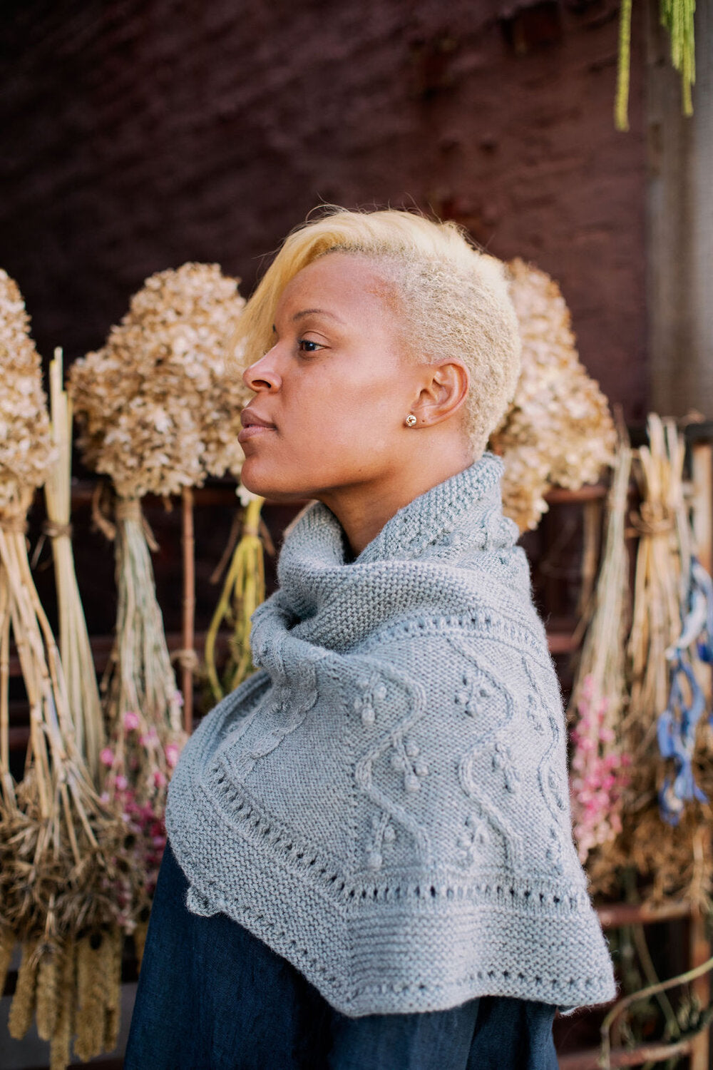 Light blue shawl knitting pattern shown on a woman in front of flower arrangements.