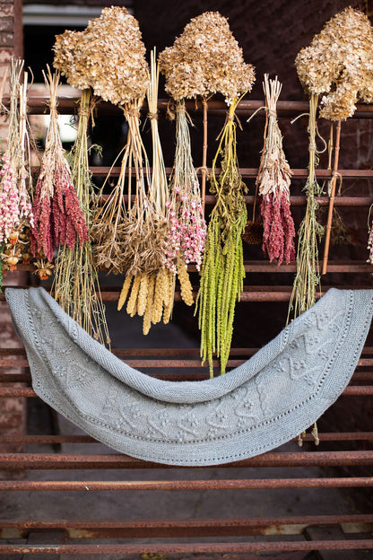 Dried floral arrangements hanging above a knitted shawl on a wooden shelf, showing the Lady of the Lake knitting pattern.