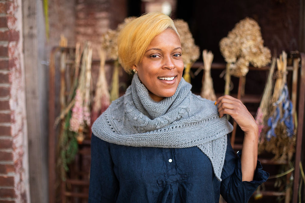 Woman wearing a gray hand-knitted scarf in front of a floral arrangement, showing the finished Lady of the Lake pattern.