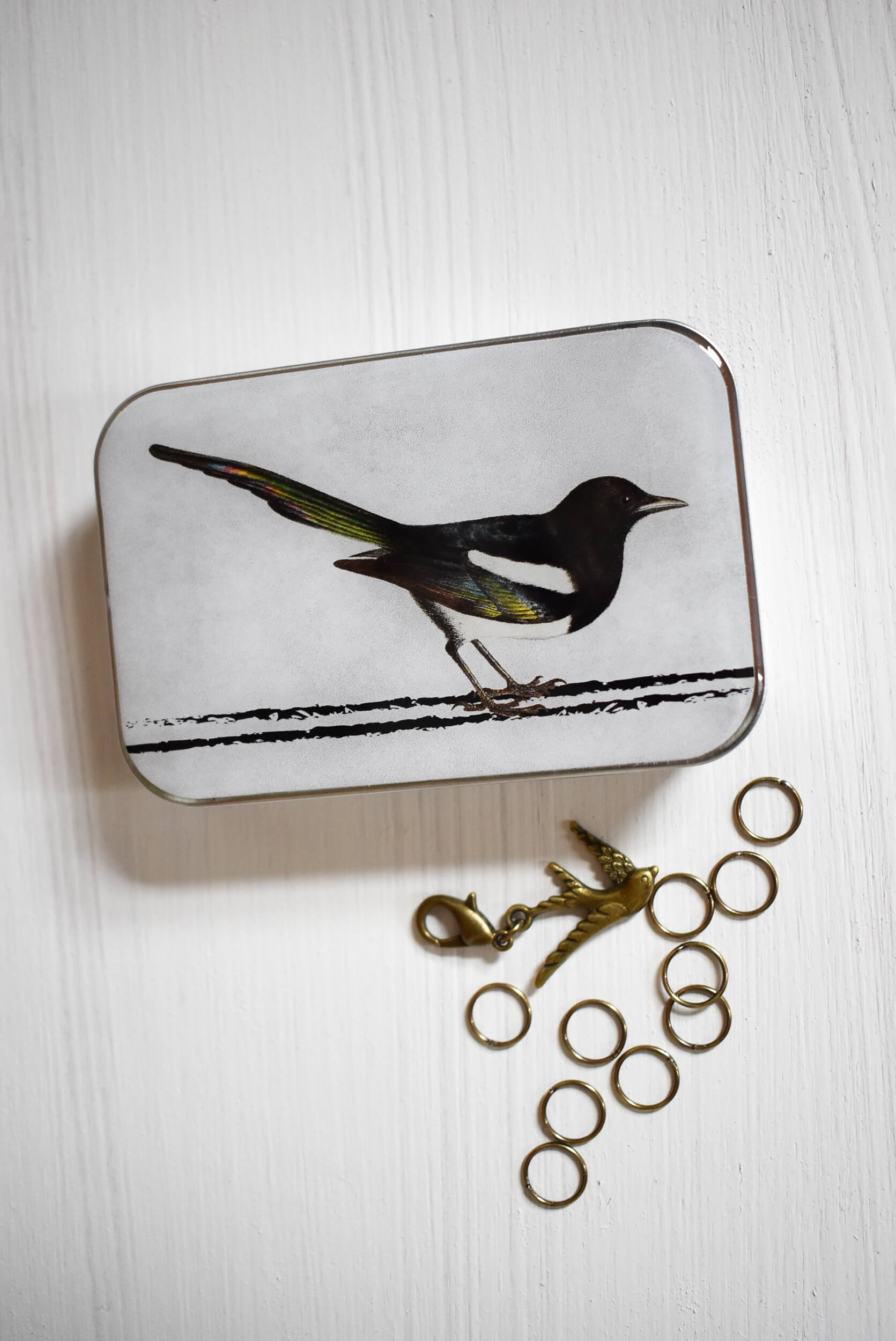 Magpie knitting notions tin with 10 round golden stitch markers and one bird progress keeper.