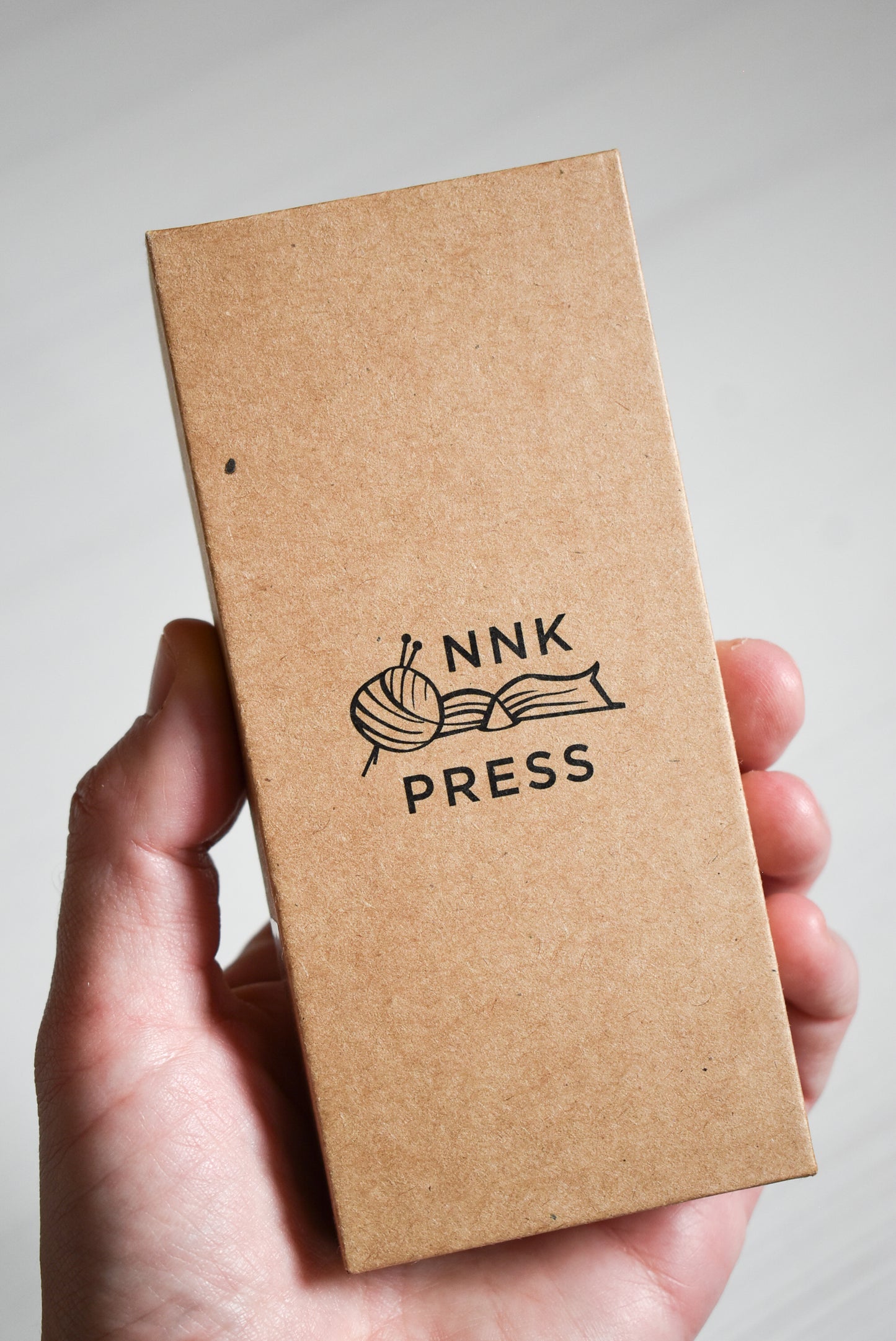Yarn scissors paper box with 'NNK Press' logo held in a hand.
