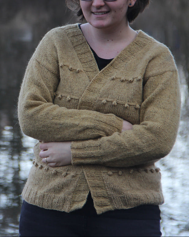 Woman wearing a mustard yellow sweater standing by a lake, wearing the finished Gooseberry Cardigan.