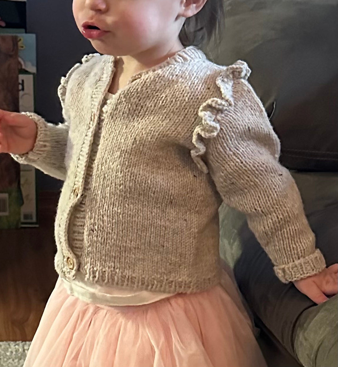 Child wearing a beige knitted cardigan based on the Ruffle Baby Cardigan knitting pattern by Katerina Rieckel.