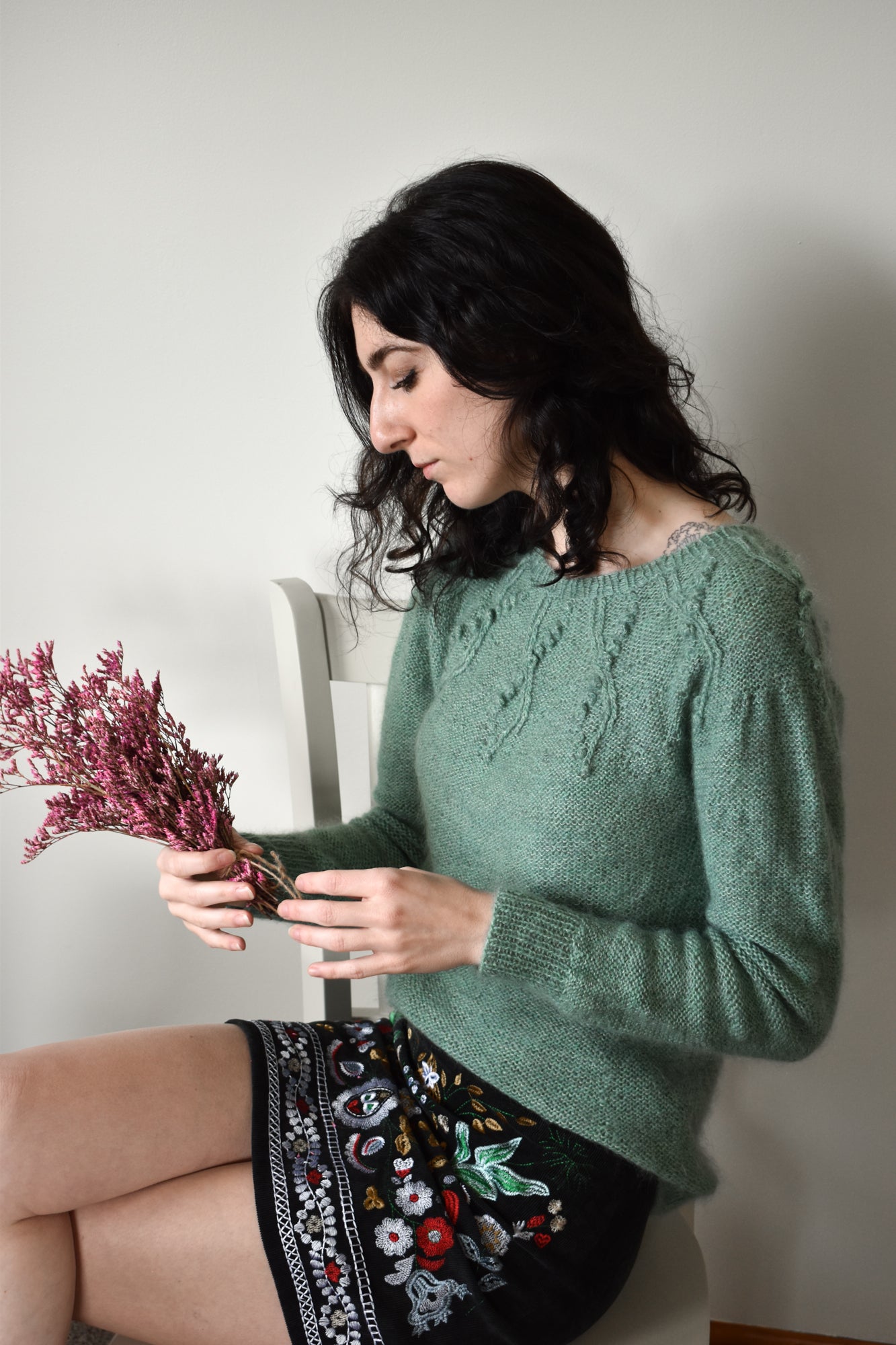 Woman in a green sweater holding a bouquet of flowers, illustrating the Seaweed Pullover knitting pattern.