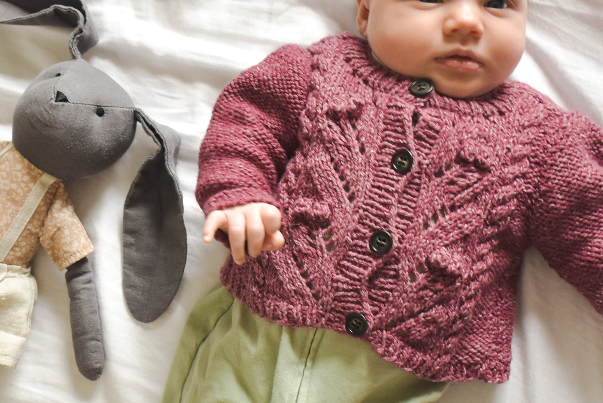 Baby wearing a knitted pink sweater based on Sofie's Cardigan knitting pattern, lying on a white blanket next to plush toys.
