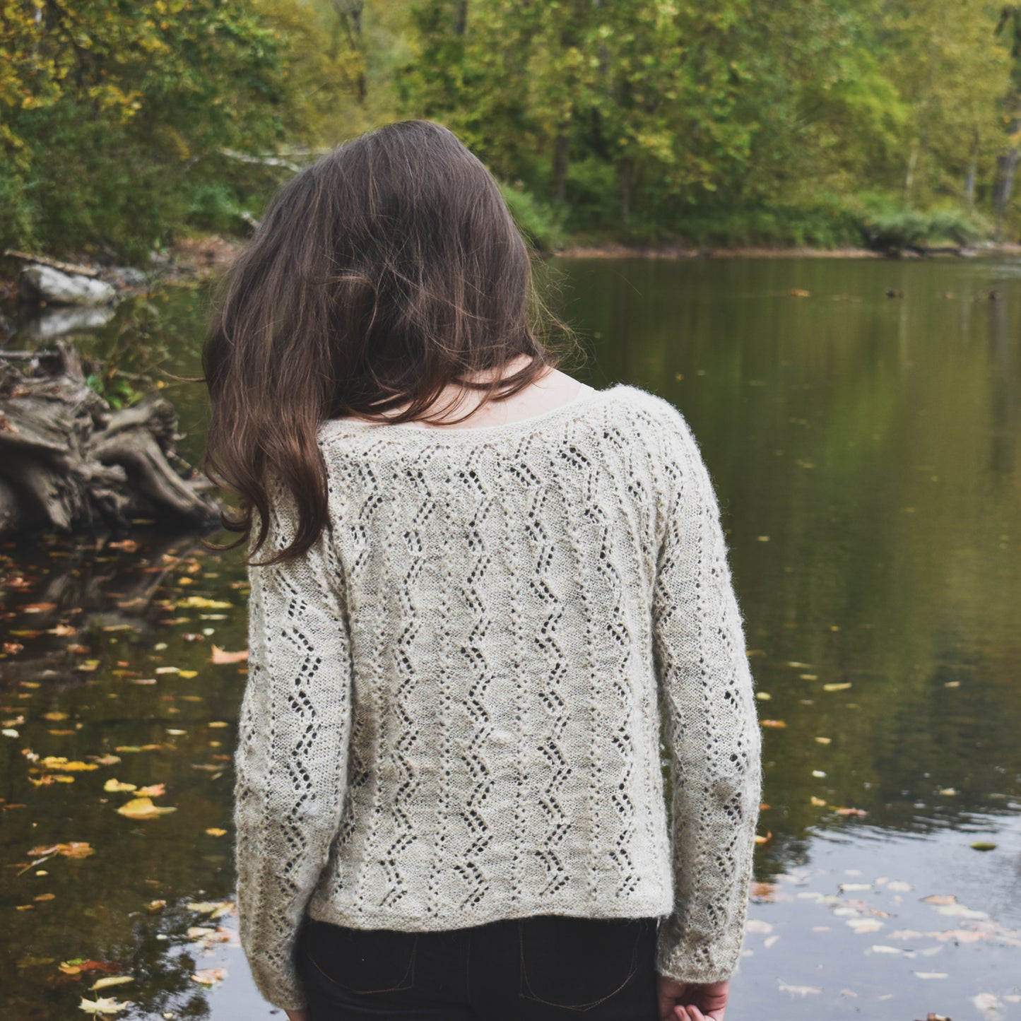 Person wearing a patterned sweater standing by a lake with trees in the background, illustrating the Battenkill Pullover knitting pattern.