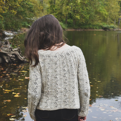 Person wearing a patterned sweater standing by a lake with trees in the background, illustrating the Battenkill Pullover knitting pattern.