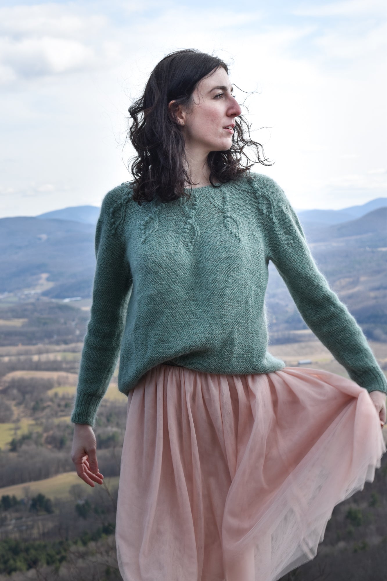 Woman wearing a green sweater based on the Seaweed Pullover knitting pattern, and pink skirt standing in a scenic outdoor setting with mountains.