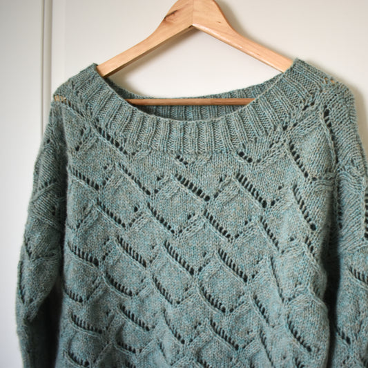 Teal hand-knit textured sweater on a wooden hanger against a white wall.