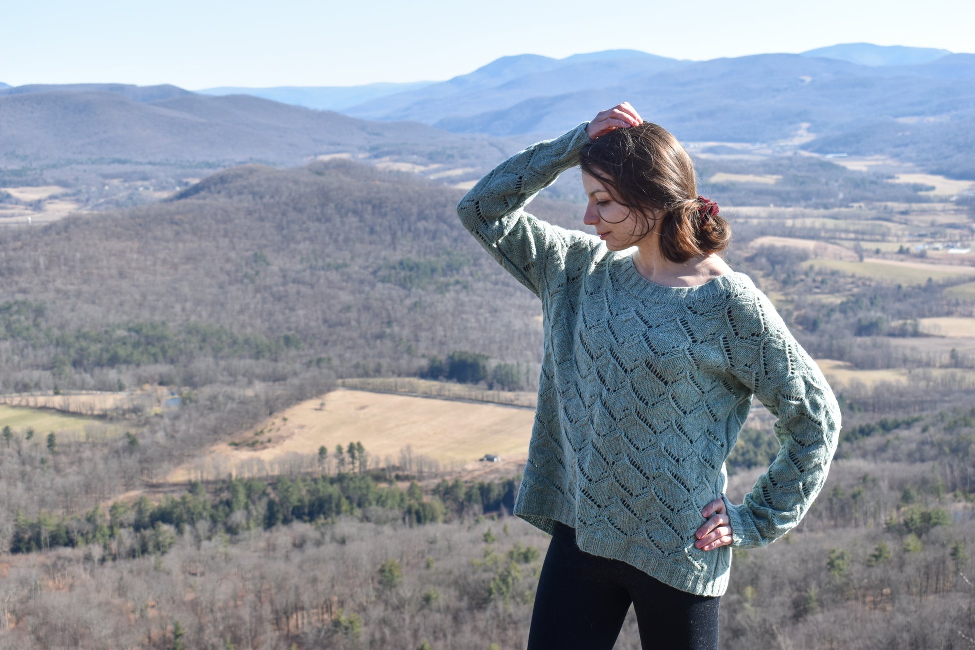 Woman wearing a green Waterscapes sweater standing in a scenic landscape with mountains and fields.