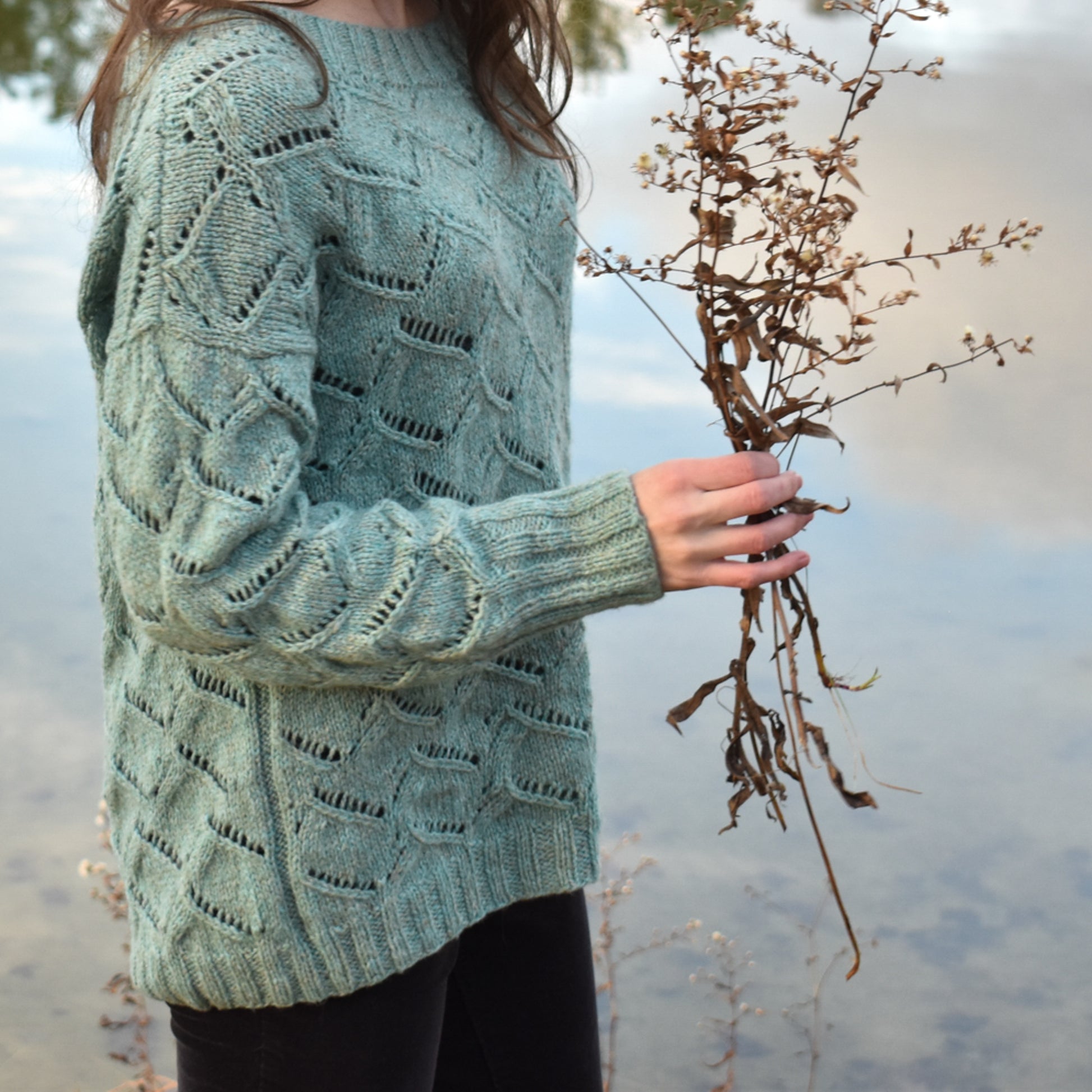 Woman wearing a textured green sweater holding dried plants by a body of water.