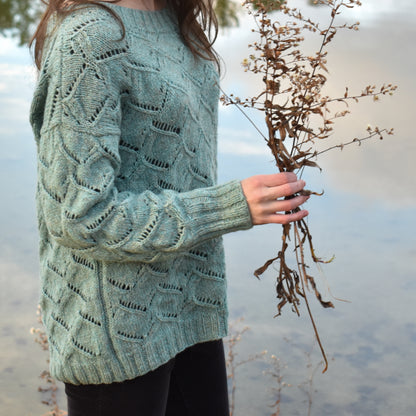 Woman wearing a textured green sweater holding dried plants by a body of water.