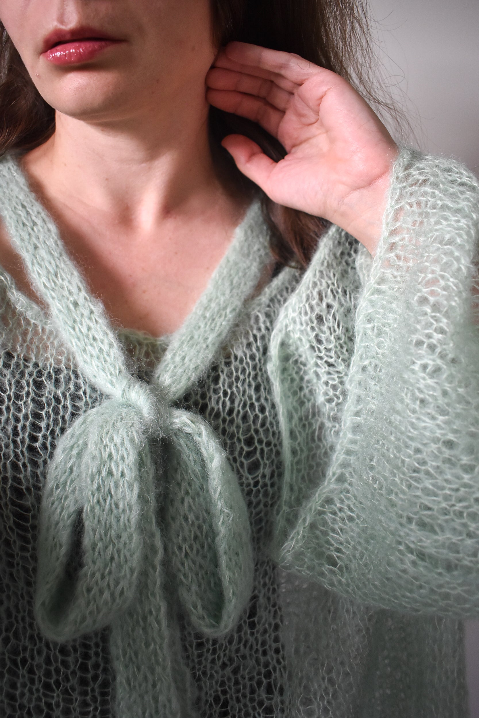 A close-up of a person wearing a pale sage green, open-knit mohair blouse knitting pattern with a tie at the neckline. The hand is touching their hair, and the background is a soft, neutral gray.