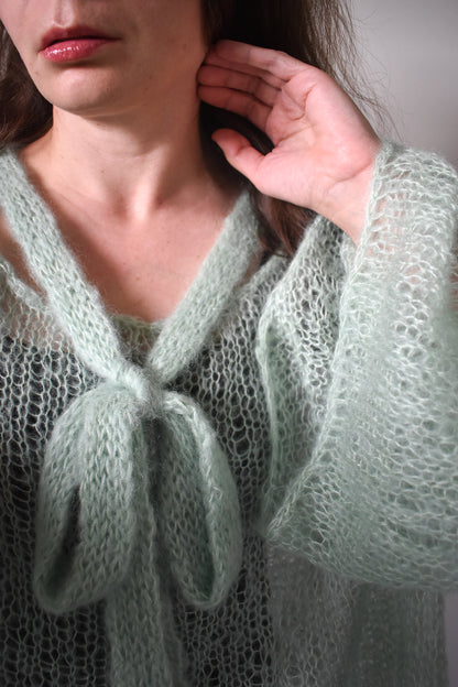 A close-up of a person wearing a pale sage green, open-knit mohair blouse knitting pattern with a tie at the neckline. The hand is touching their hair, and the background is a soft, neutral gray.