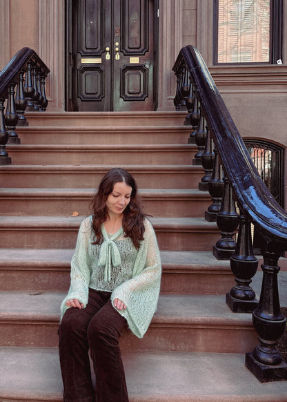 A woman sits on the steps of a brownstone building, wearing a light sage green knitted blouse based on a PDF pattern.