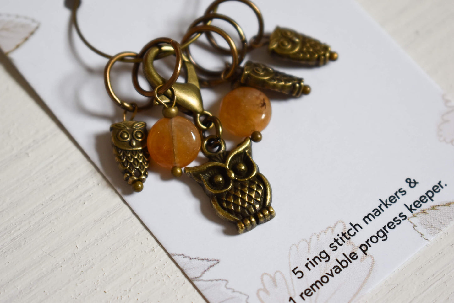 Bronze owl-themed stitch markers with orange beads on a card.