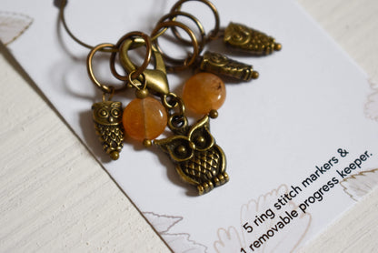 Bronze owl-themed stitch markers with orange beads on a card.
