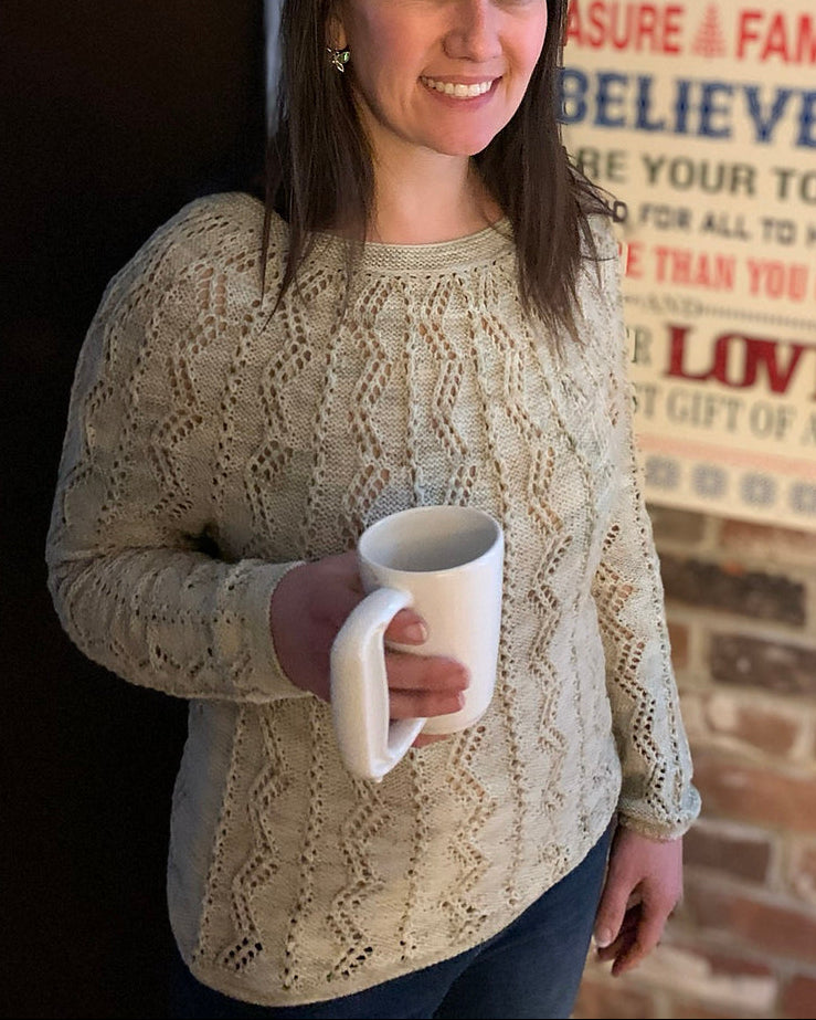 Woman holding a mug in front of a brick wall with a Christmas-themed sign, wearing a finished Battenkill Pullover.